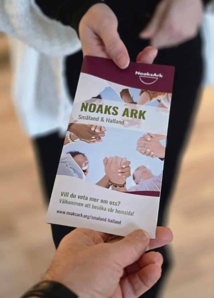 En hand som räcker över en broschyr om Noaks Ark Småland och Halland till en annan hand